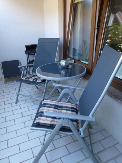 Ein gemütlicher Balkon mit zwei Stühlen und einem Tisch. Der Tisch hat eine kleine Dekoration in der Mitte und große Fenster im Hintergrund.
