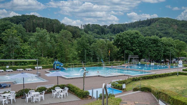 Ein schönes Freibad mit mehreren Becken und einer grünen Umgebung. Im Hintergrund sind Bäume und ein blauer Himmel zu sehen.