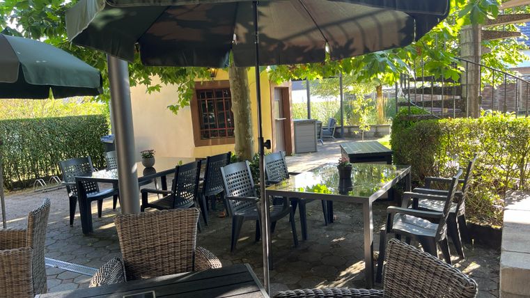 Ein einladender Außenbereich mit Tischen und Stühlen unter Sonnenschirmen. Umgeben von grünen Pflanzen und einer ruhigen Atmosphäre.