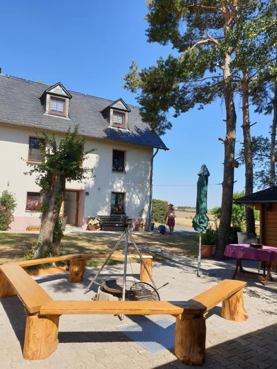 Un espace extérieur confortable avec un foyer en bois, entouré d'arbres. En arrière-plan, se trouve une belle maison sous un ciel bleu.