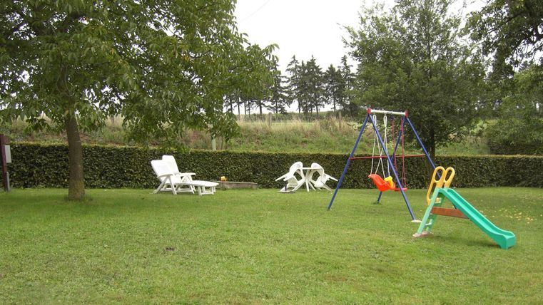 Un jardin vert avec une aire de jeux, qui comprend une balançoire et un toboggan. À l'arrière-plan, il y a des chaises longues sous un arbre.