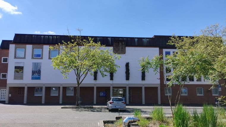 Ein modernes Gebäude mit großen Fenstern und einem klaren blauen Himmel. Im Vordergrund sind Bäume und eine Parkmöglichkeit zu sehen.