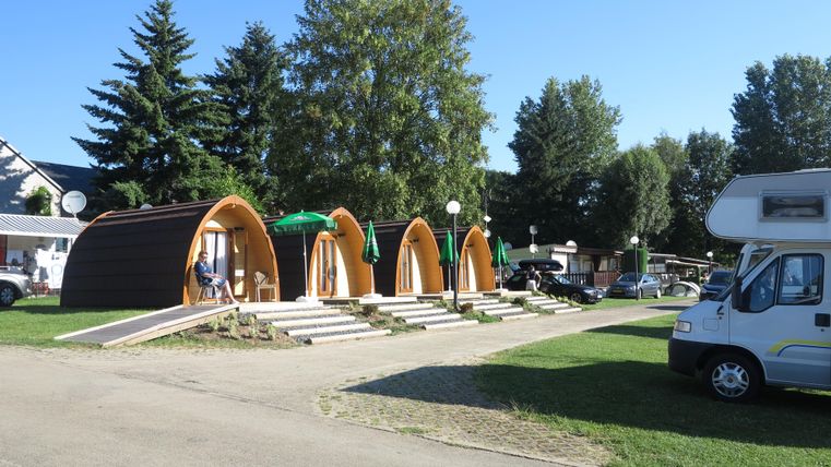 Moderne Unterkünfte in Form von Hütten stehen in einer grünen Umgebung. Im Hintergrund sind Bäume und Wohnmobile sichtbar.