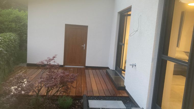 Een moderne huisaanzicht met een klein houten dek en een voordeur. Op de voorgrond zijn enkele planten en het pad van tegels te zien.