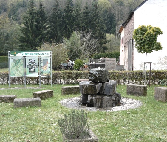 Ein Brunnen aus Steinen in Mettendorf, umgeben von Bänken. Im Hintergrund eine Infotafel des Naturparks Südeifel und ein Gebäude., © Felsenland Südeifel Tourismus, Christian Calonec-Rauchfuss