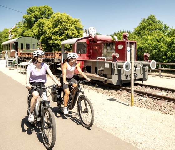Zwei Radfahrerinnen auf einem Radweg neben einer alten Lokomotive im Eisenbahnmuseum Pronsfeld, umgeben von grünen Bäumen., © Eifel Tourismus GmbH, Dominik Ketz