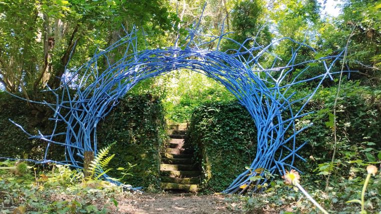 Ein bunter Torbogen aus blauen Zweigen führt in einen grünen Garten. Der Weg ist von Pflanzen und Bäumen umgeben.