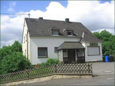 Ein einfaches, zweigeschossiges Haus mit einem grauen Dach. Vor dem Haus befindet sich ein Zaun und einige grünen Pflanzen.