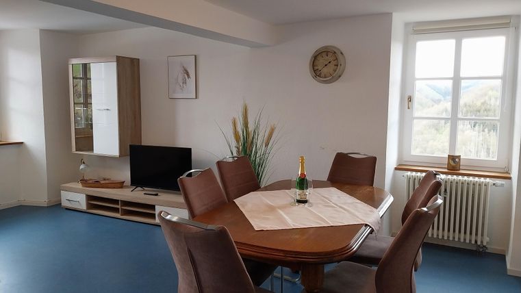 Ein heller Raum mit einem Essbereich und einem Tisch für sechs Personen. Im Hintergrund ist ein Fernseher und ein Fenster mit Ausblick.