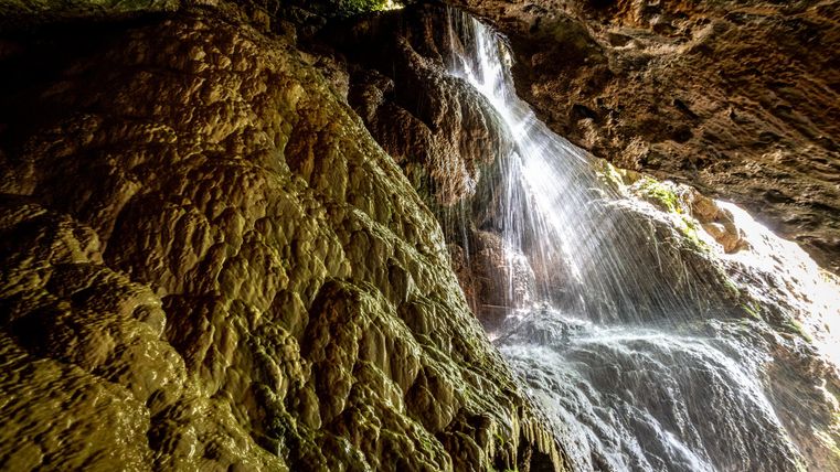 Ein Wasserfall fließt über eine steinige Felswand in einer dunklen Höhle. Das Licht reflektiert sich auf dem Wasser und sorgt für eine geheimnisvolle Atmosphäre.