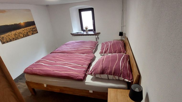 Een gezellige slaapkamer met een tweepersoonsbed, rode dekens en een klein nachttafeltje. Aan de muur hangt een schilderij van een landschap bij zonsondergang.