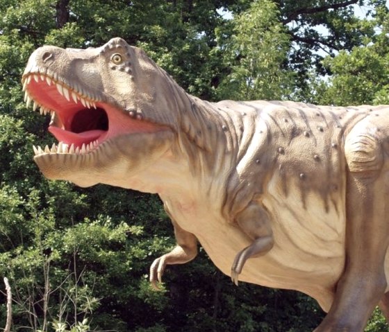 A life-size model of a Tyrannosaurus Rex stands on a rock, surrounded by trees in the background.