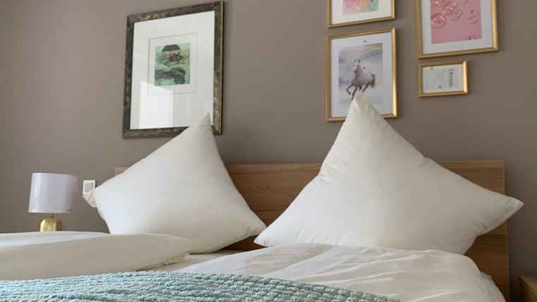 Een gezellige slaapkamer met een houten bed en grote kussens. Aan de muur hangen decoratieve afbeeldingen in lijsten en een nachtkastje met een lamp is zichtbaar.