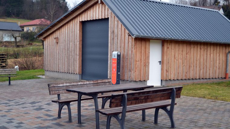 Ein Holzgebäude mit einem großen Rollladen und einem weißen Tor. Vor dem Gebäude stehen zwei Holztische und Bänke auf einem gepflasterten Bereich.