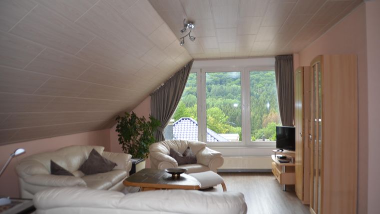A cozy living room with white furniture and large windows. The view looks out onto a green landscape.