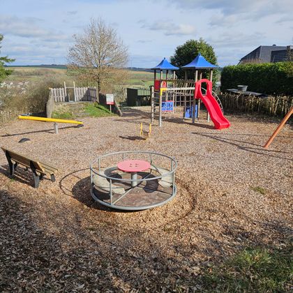 Een speelplaats met een glijbaan, een klimrek en een draaiplatform. De bodem is bedekt met houtsnippers en er zijn enkele bomen en een uitzicht op het landschap.