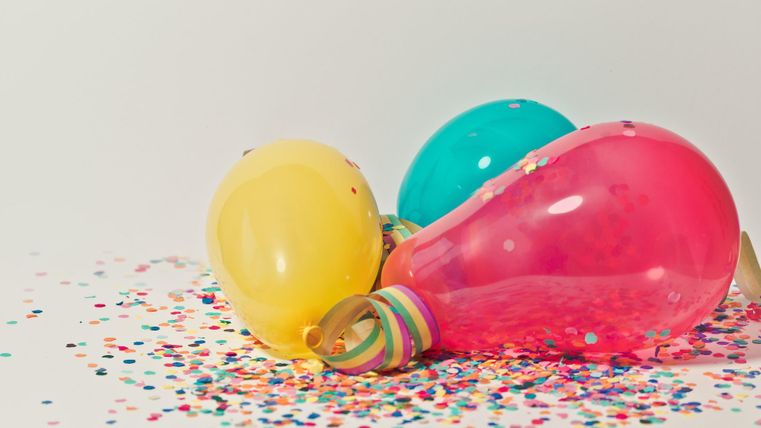 Bunte Luftballons in Gelb, Blau und Pink liegen auf einem bunten Konfettihaufen. Die Szene strahlt eine festliche Stimmung aus.
