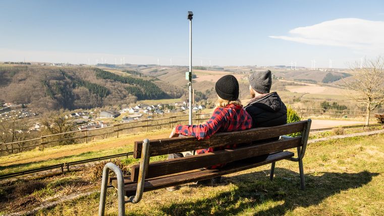 Ein Paar sitzt auf einer Bank und schaut auf die weite Landschaft. Im Hintergrund sind Hügel und Windräder zu sehen.