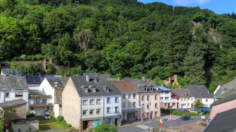 Eine kleine Stadt mit mehreren Häusern steht am Fuße eines grünen Hügels. Der blaue Himmel und die Bäume verleihen der Umgebung eine ruhige Atmosphäre.