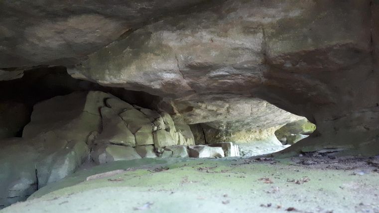 Une grotte naturelle avec de grandes roches grises. Le sol est sablonneux et il y a peu de lumière.