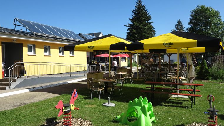 Eine gemütliche Terrasse mit sonnigen Gelben Sonnenschirmen und Tischen. Im Hintergrund ist ein gelbes Gebäude und ein grüner Garten zu sehen.