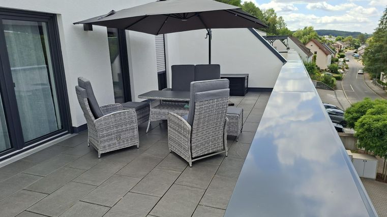 Une terrasse moderne avec une table à manger et des chaises en rotin. Un grand parasol procure de l'ombre et à l'arrière-plan, on peut voir quelques maisons.