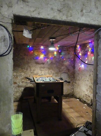 Ein einfacher Raum mit einem Tischfußballspiel und bunten Lichtern an der Wand. Der Raum hat eine rustikale Atmosphäre mit Steinen und Holz.