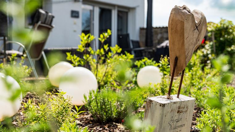 Eine Holzskulptur steht in einem Garten, umgeben von bunten Pflanzen und wenigen leuchtenden Ballons. Im Hintergrund ist ein Haus mit großen Fenstern zu sehen.