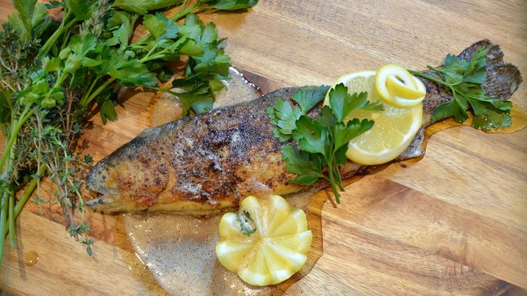 Ein gegrillter Fisch liegt auf einem Holzbrett, umgeben von frischen Kräutern. Neben dem Fisch sind Zitronenscheiben und eine dekorative Zitronenblüte platziert.