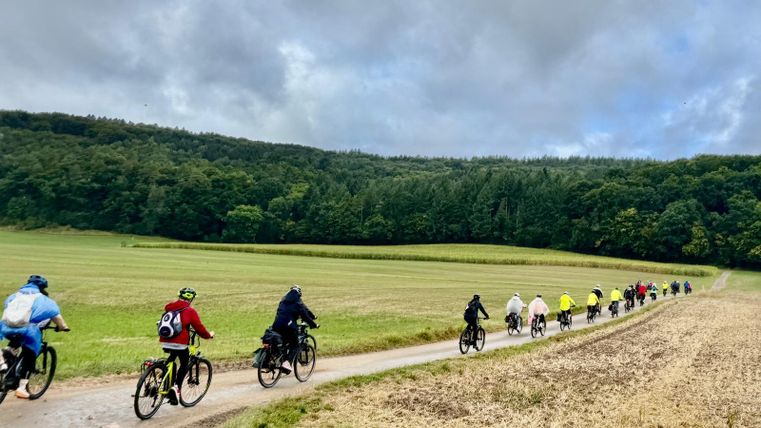 Vier Personen stehen vor einem Café und posieren mit einem E-Bike. Sie lächeln und tragen sportliche Kleidung.