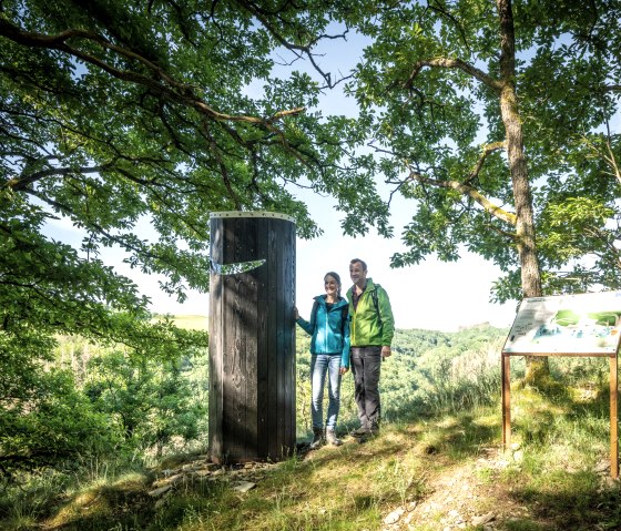 Two people are standing next to a wooden pillar in the forest. An information board is on the right of the picture. The surroundings are green and sunny., © Eifel Tourismus GmbH, Dominik Ketz