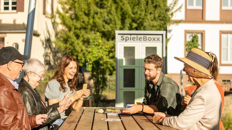 Eine Gruppe von Menschen spielt Karten an einem Tisch im Freien. Im Hintergrund ist eine Spielebox zu sehen, umgeben von Bäumen und Gebäuden.