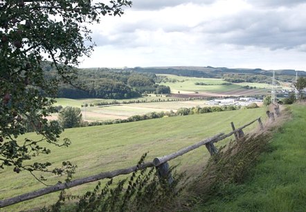 Landschaft mit Wiesen, B&auml;umen und einem Holzzaun im Vordergrund. Im Hintergrund sind Felder und H&uuml;gel unter einem bew&ouml;lkten Himmel zu sehen., &copy; V. Teuschler