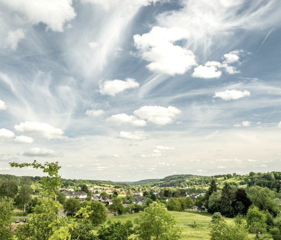 Green landscape with fields, trees and a village. The sky is blue with white clouds stretching across the scene., © Eifel Tourismus, Dominik Ketz