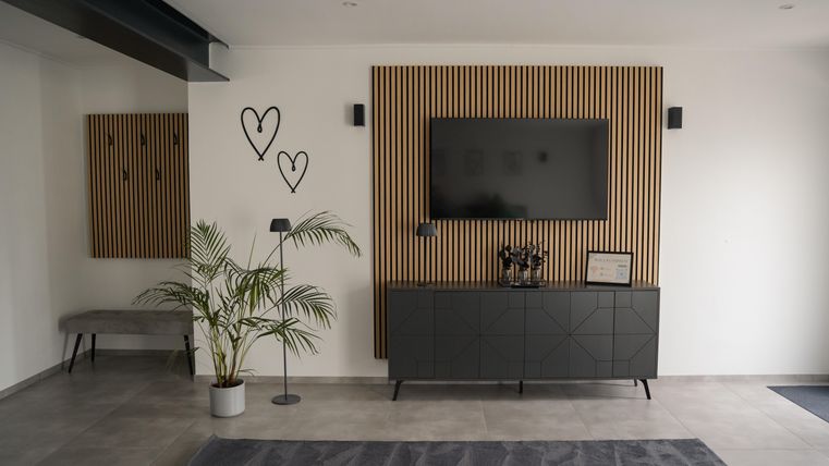 A modern living room with wood paneling and a TV mounted on the wall. A plant and a black sideboard complement the simple, elegant decor.