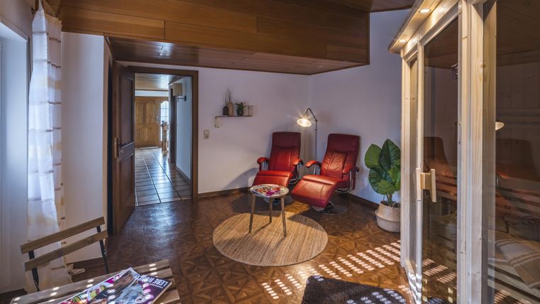 Ein gemütlicher Raum mit zwei roten Sesseln, einem runden Tisch und einem großen Pflanzen. Helle Farben und sanftes Licht sorgen für eine einladende Atmosphäre.