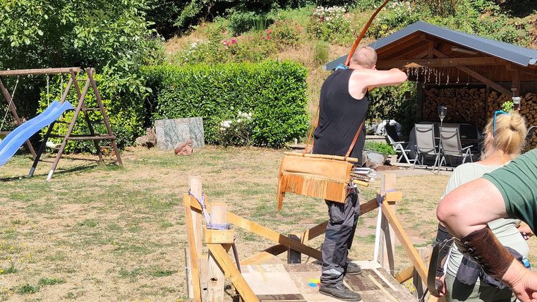 Ein Mann schießt mit einem Bogen auf einem Holzstand. Im Hintergrund sieht man eine Gartenlandschaft mit einer Schaukel und einem überdachten Sitzbereich.