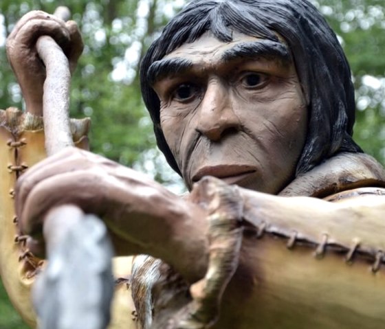 Prehistoric figure with spear in Dinopark, surrounded by trees. Audio tour interface visible.