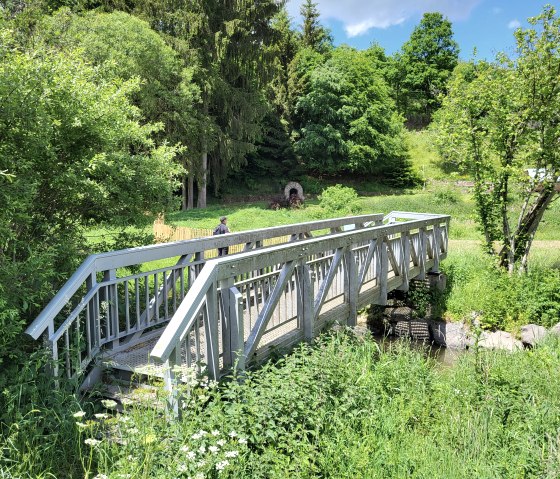 Eine kleine Holzbr&uuml;cke f&uuml;hrt &uuml;ber einen Bach in einer gr&uuml;nen, bewaldeten Landschaft. Ein Spazierg&auml;nger ist im Hintergrund zu sehen., &copy; Tourist-Information Islek, Ingrid Wirtzfeld