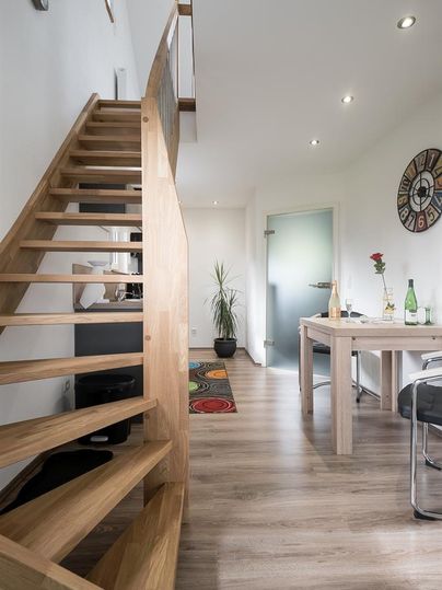 Un escalier moderne en bois mène à une pièce lumineuse. Au premier plan se trouve une table avec quelques plantes et des boissons.