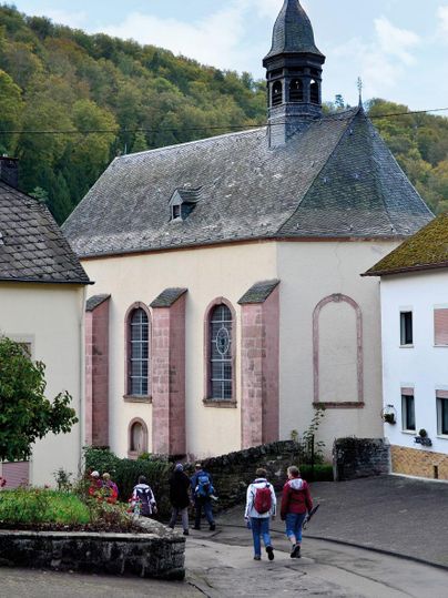 Eine kleine Kirche mit einer spitzen Spitze und bunten Fenstern in einer ruhigen Straße. Wanderer gehen entspannt vorbei, umgeben von grünen Hügeln.