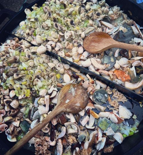 Eine große Pfanne mit einer Mischung aus frischen Meeresfrüchten, Gemüse und Kräutern. Zwei Holzlöffel liegen zum Rühren bereit.