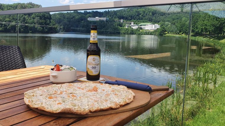 A table with a large pizza, a bottle of beer, and a small salad. In the background, a picturesque lake and green countryside can be seen.
