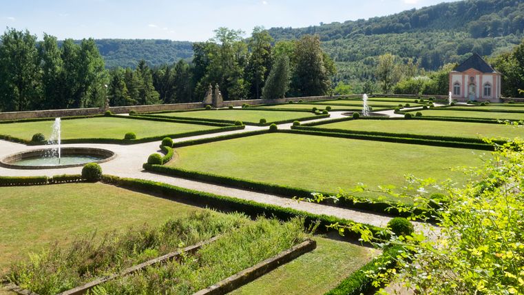 Französischer Garten mit Springbrunnen und Pavillon im Hintergrund, umgeben von Bäumen und Hügeln.