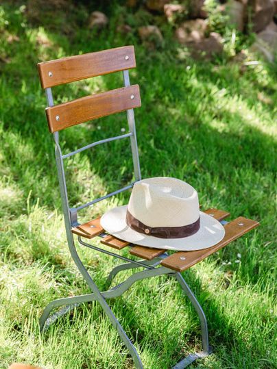 Ein eleganter Stuhl aus Metall und Holz steht auf grünem Gras. Darauf liegt ein helles Hut mit einem braunen Band.
