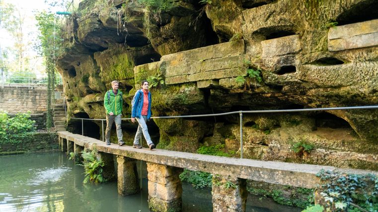 Zwei Personen gehen auf einem Steg entlang einer Felswand in einer grünen Landschaft. Im Vordergrund ist ein ruhiges Wasser zu sehen, umgeben von üppiger Vegetation.