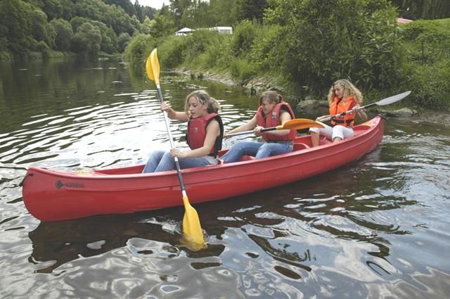 Eine Gruppe von drei Frauen paddelt in einem roten Kayak auf einem ruhigen Fluss. Umgeben von grüner Natur genießen sie einen aktiven Tag im Freien.