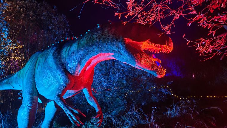 Ein leuchtender Dinosaurier steht in einem dunklen Umfeld, umgeben von bunten Lichtern. Die Farben sind intensiv und verleihen der Szene eine aufregende Atmosphäre.