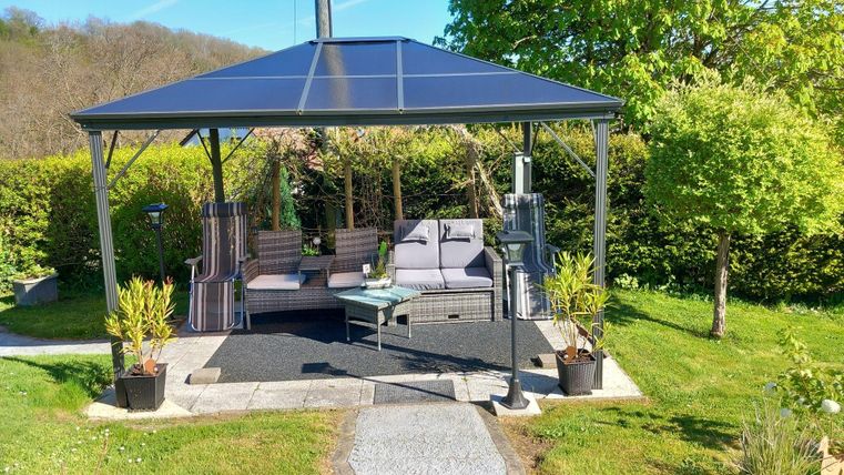 A cozy garden pavilion with seating and a table. Surrounded by lush greenery and pretty plants.