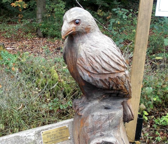 A wooden sculpture of a bird sits on a tree trunk in the forest. A plaque is attached to the sculpture., &copy; Tourist-Information Bitburger Land